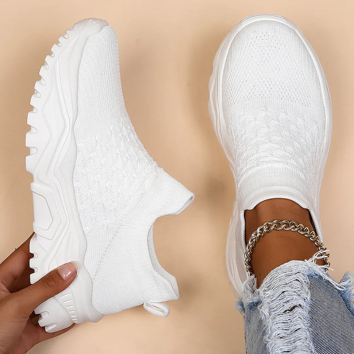 Pair of white sock sneakers with chunky soles, one worn with frayed jeans and gold anklet on beige background
