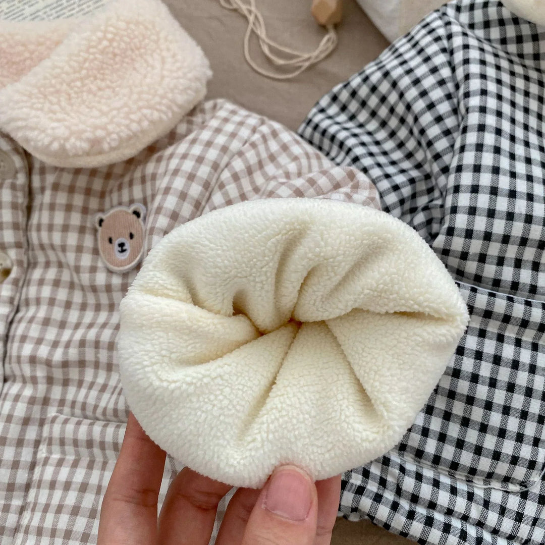 Hand holding soft cream sherpa fabric pom pom on checked beige and black baby clothes with bear patch