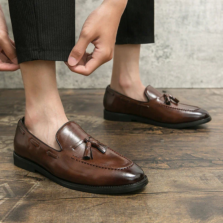 Brown leather tassel loafers worn with black pants on wooden floor and gray wall background
