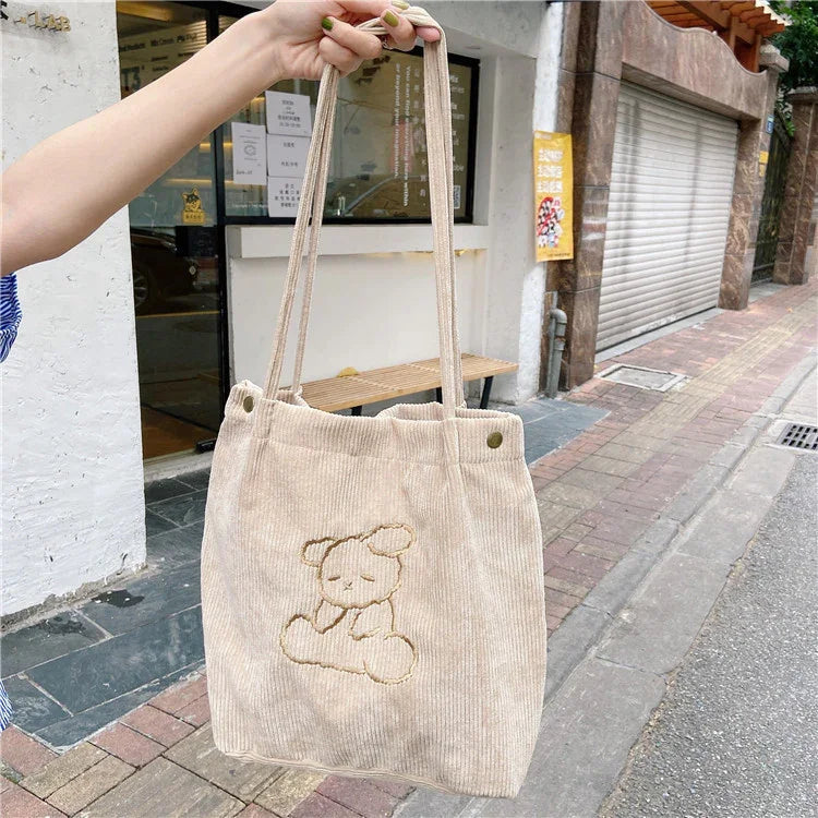 Beige corduroy tote bag with embroidered sleeping bear design held outside near sidewalk and storefront