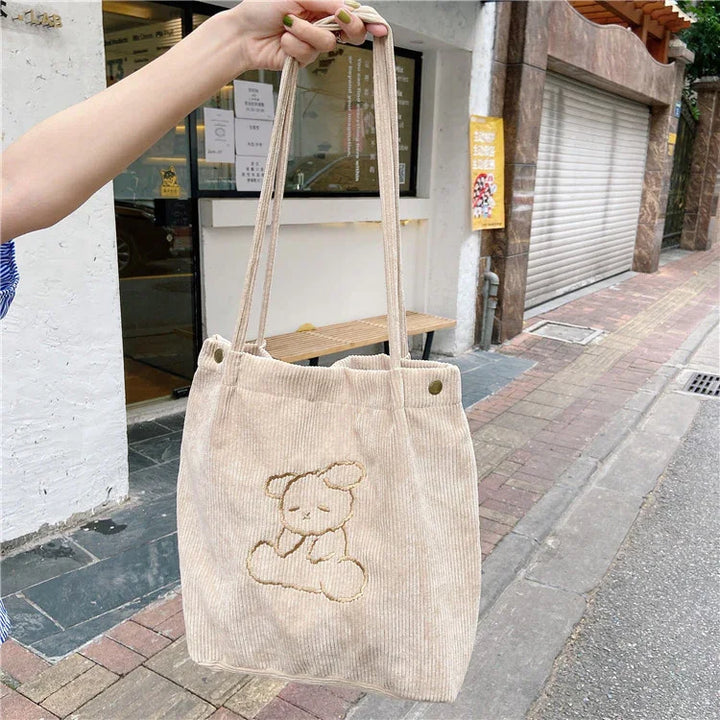 Beige corduroy tote bag with embroidered sleeping bear design held outside near sidewalk and storefront