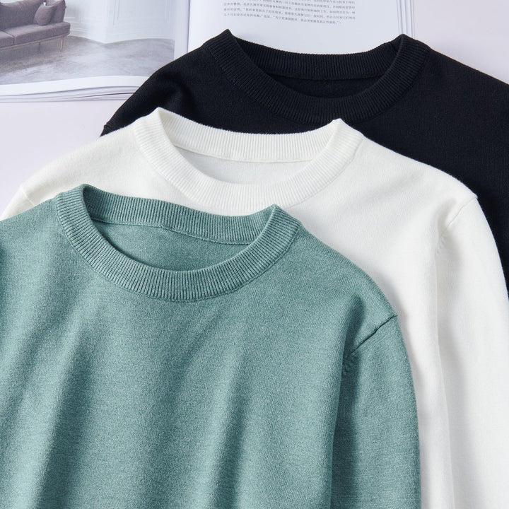 Stacked green, white, and black crewneck sweaters on a white surface with an open book background