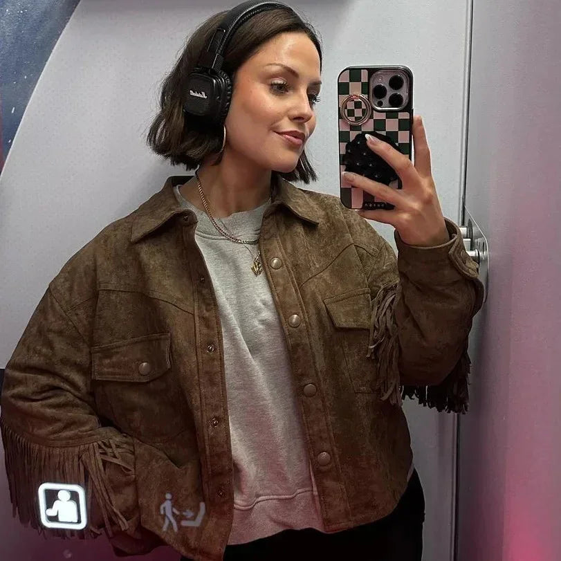 Woman wearing brown fringe jacket and gray sweatshirt taking mirror selfie with headphones and checkered phone case