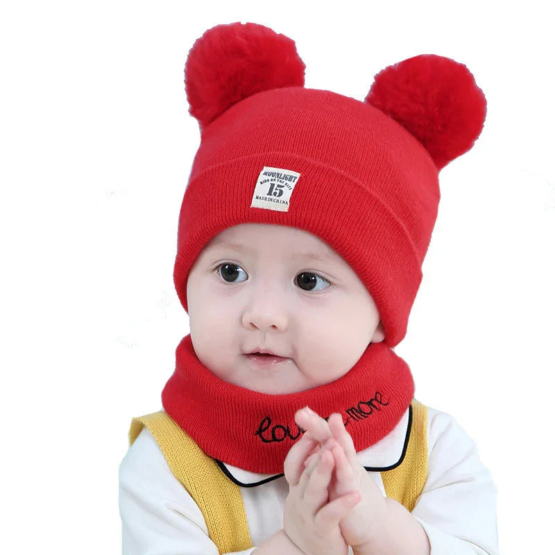 Baby wearing a red knit hat with two pom-poms and matching neck warmer, yellow suspenders, white shirt