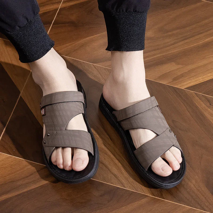 Person wearing brown cross-strap sandals with black soles on wooden floor