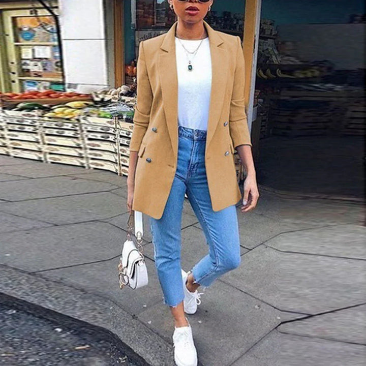 Woman wearing tan blazer, white top, blue jeans, white sneakers, and carrying white handbag outdoors