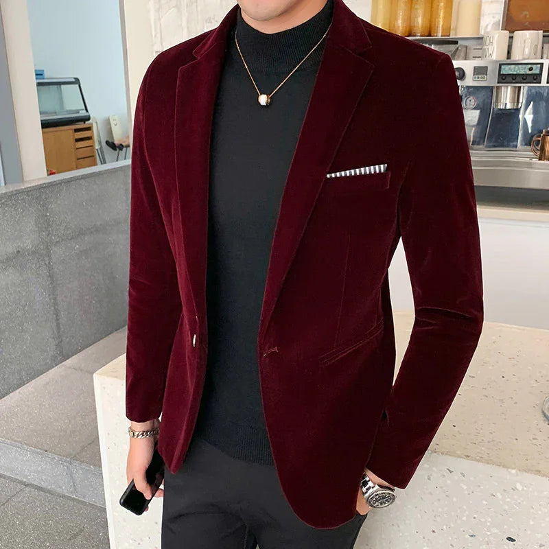 Man wearing a burgundy velvet blazer with a black turtleneck and gold necklace, holding a phone indoors
