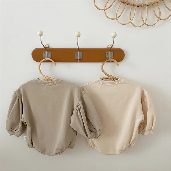 Two neutral-toned baby puff sleeve tops hanging on wooden hangers on wall hooks