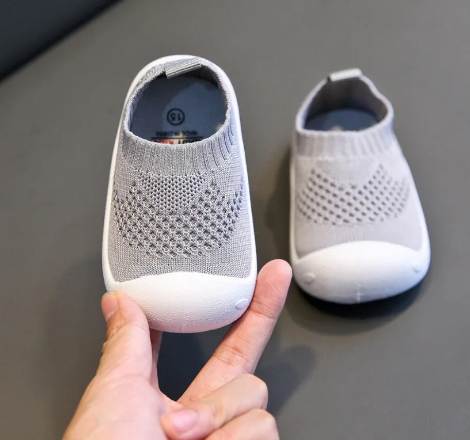 Pair of gray knitted baby shoes with white rubber soles on black surface, one held by hand