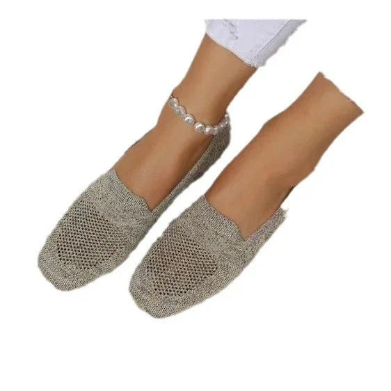 Feet wearing beige knitted slip-on shoes and white pants, with a pearl anklet