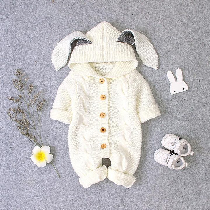 Cream knitted baby romper with bunny ears hood, wooden buttons, and white baby shoes on gray background