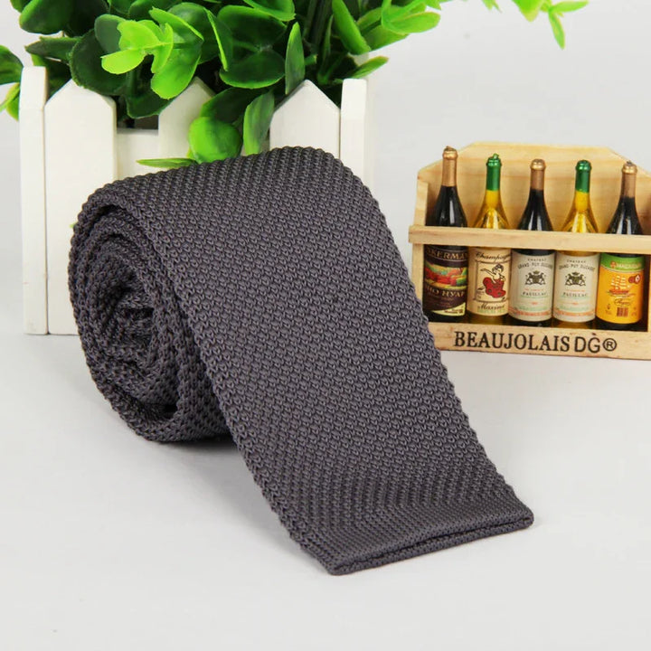 Dark gray knitted tie rolled on white surface with small wine bottle crate and green plant background