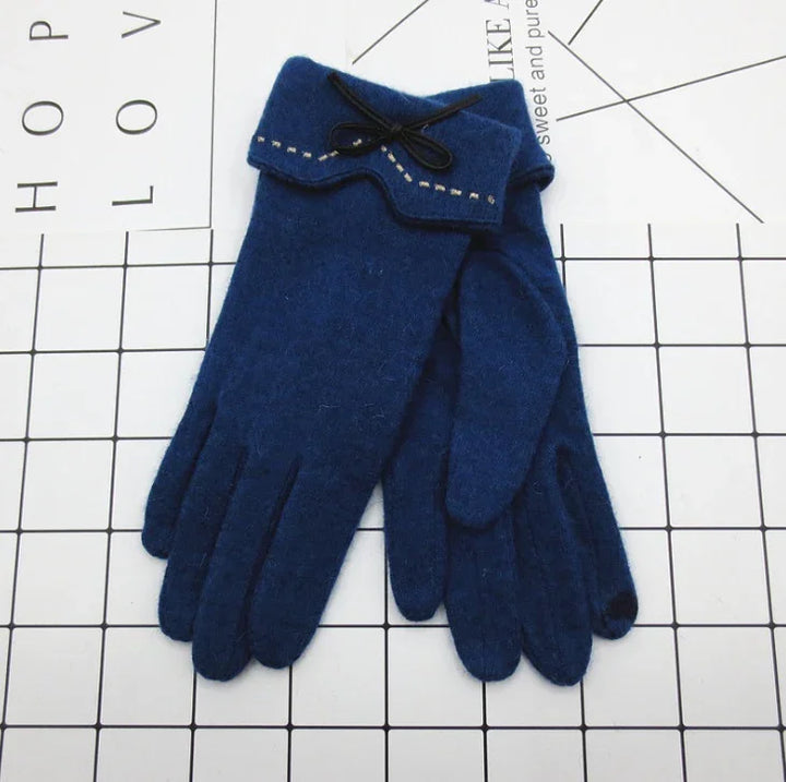 Pair of navy blue wool gloves with decorative bow and stitching detail on cuff, laid on grid background