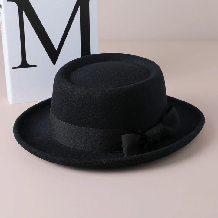 Black felt bowler hat with a ribbon detail on beige surface, white block with letter M background