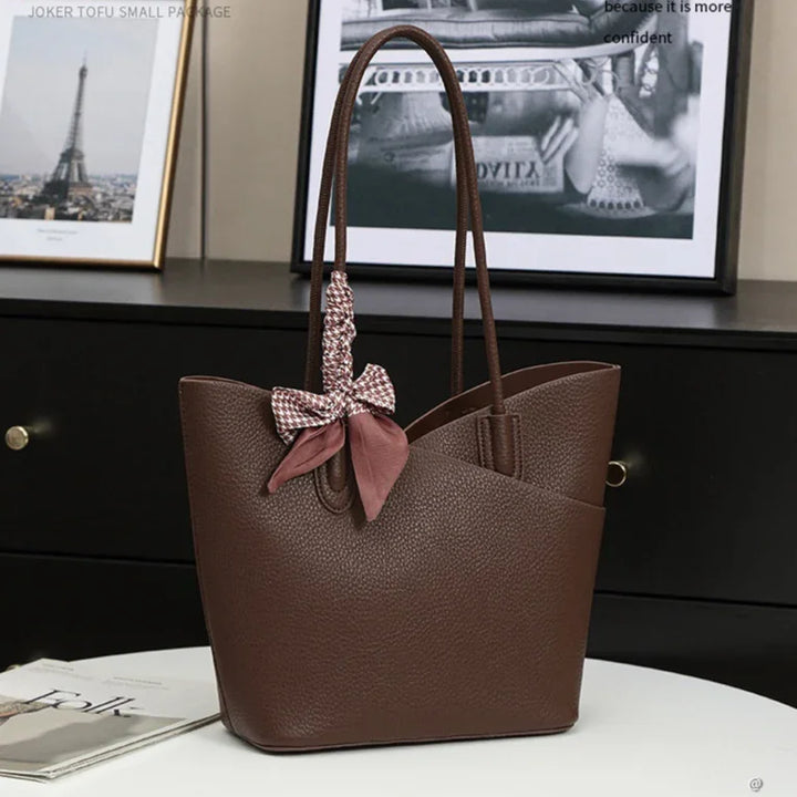 Brown textured leather tote bag with long handles and decorative patterned bow on a white table