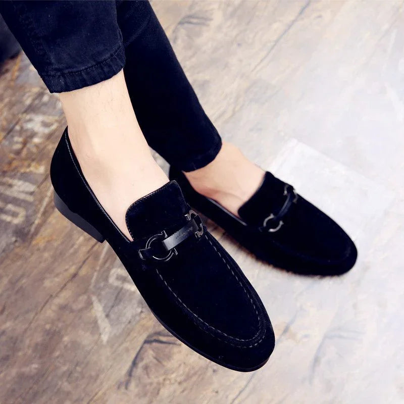 Black suede loafers with metal buckle detail worn with black pants on wooden floor