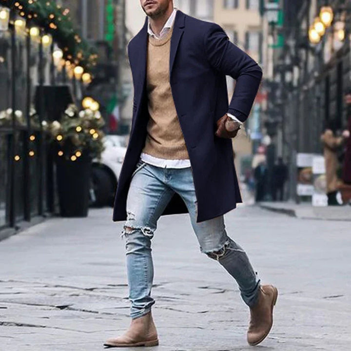 Man wearing navy blue overcoat, tan sweater, ripped light jeans, and brown boots walking on urban street