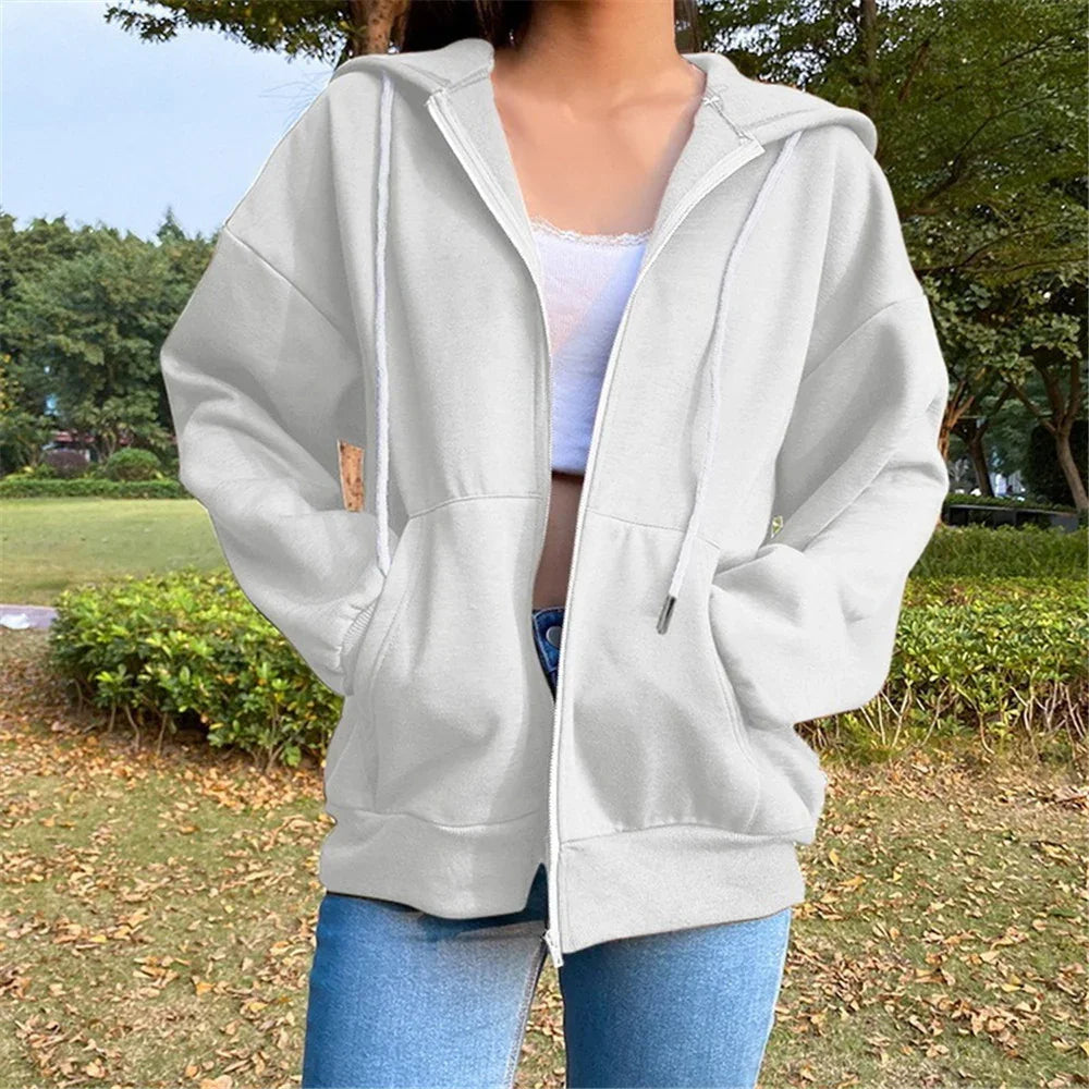 Woman wearing a white oversized zip-up hoodie and light blue jeans in a park setting with green bushes and trees