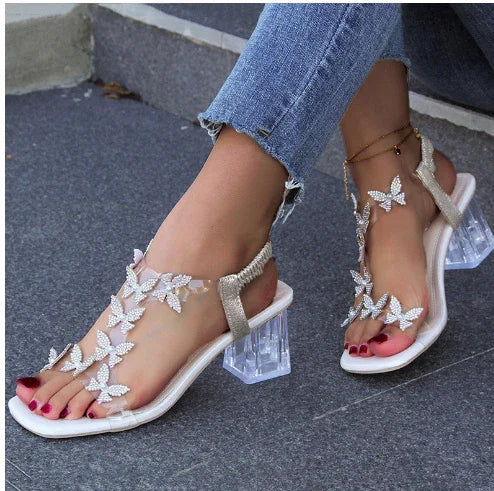 Woman wearing white butterfly embellished clear block heel sandals and ankle bracelets with red pedicure on gray pavement