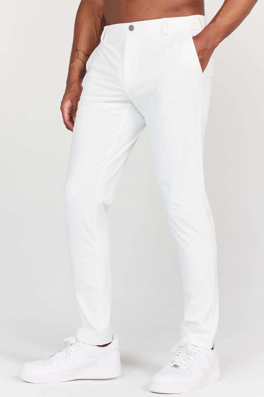 Man wearing slim fit white pants and white sneakers, with hand in pocket and visible arm tattoo