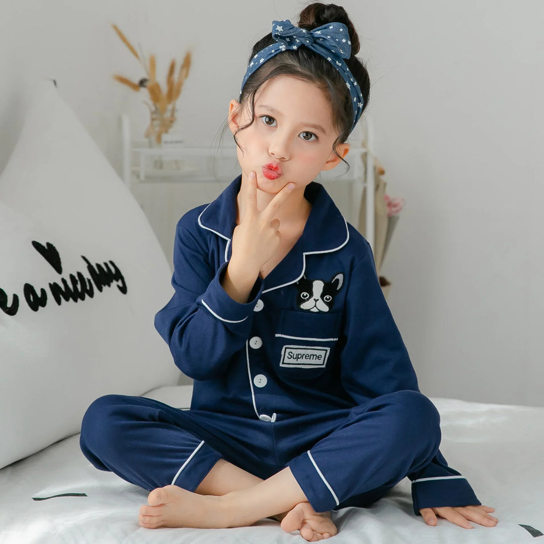 Child in navy blue pajama set with dog patch and Supreme logo, sitting on white bed with blue star headband