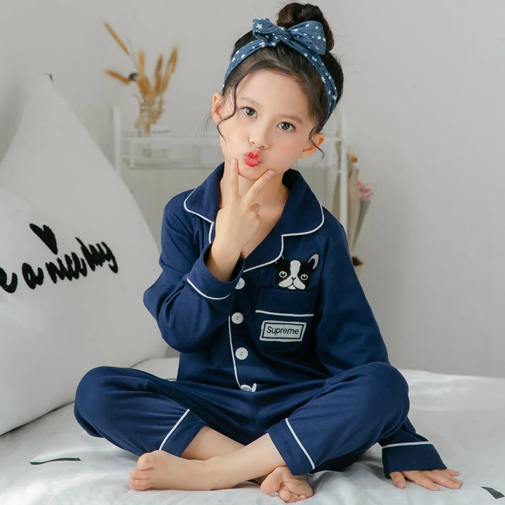 Child in navy blue pajama set with dog patch and Supreme logo, sitting on white bed with blue star headband