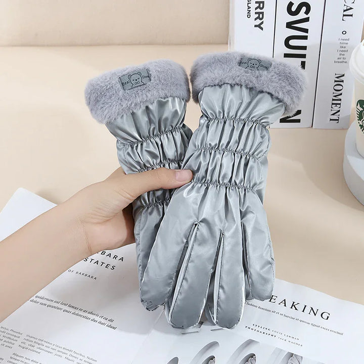 Hand holding shiny silver winter gloves with fluffy grey cuffs on a beige surface with books and papers