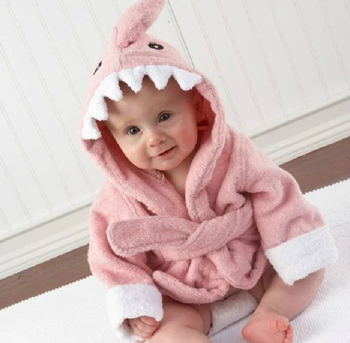 Smiling baby wearing a pink shark hooded bathrobe with white teeth detail sitting on a white rug indoors