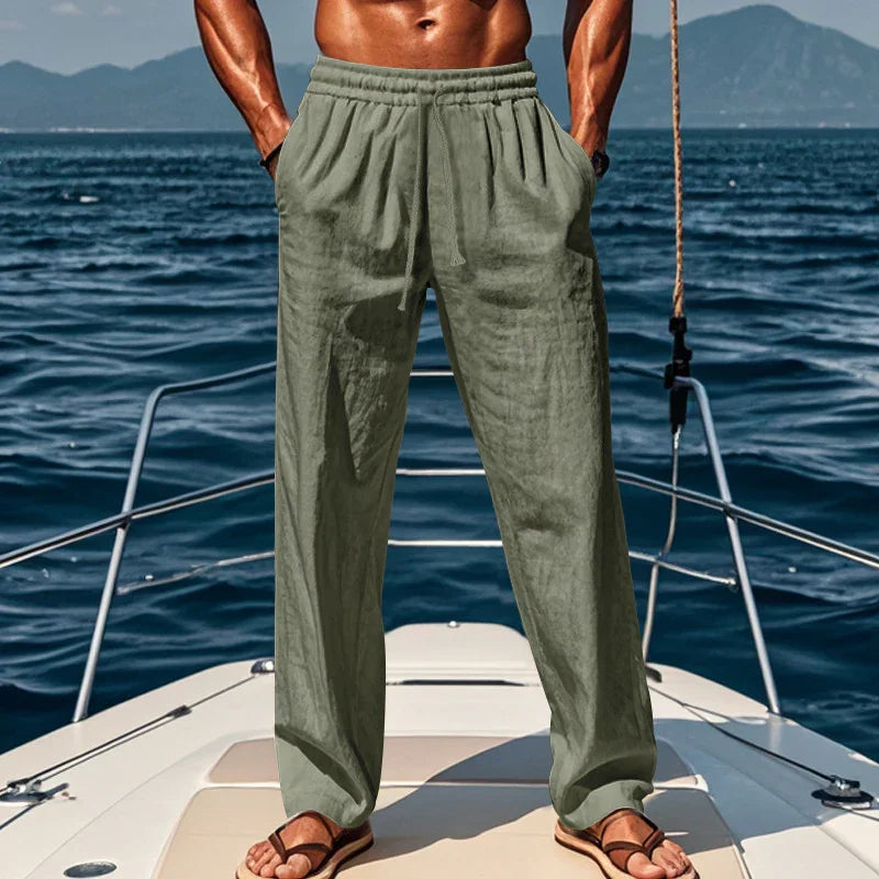 Muscular man wearing olive green linen pants and brown sandals standing on yacht deck with ocean background