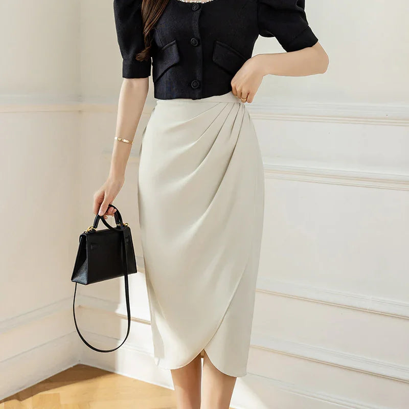Woman wearing a black cropped blouse and cream pleated midi skirt holding a black handbag in a bright room
