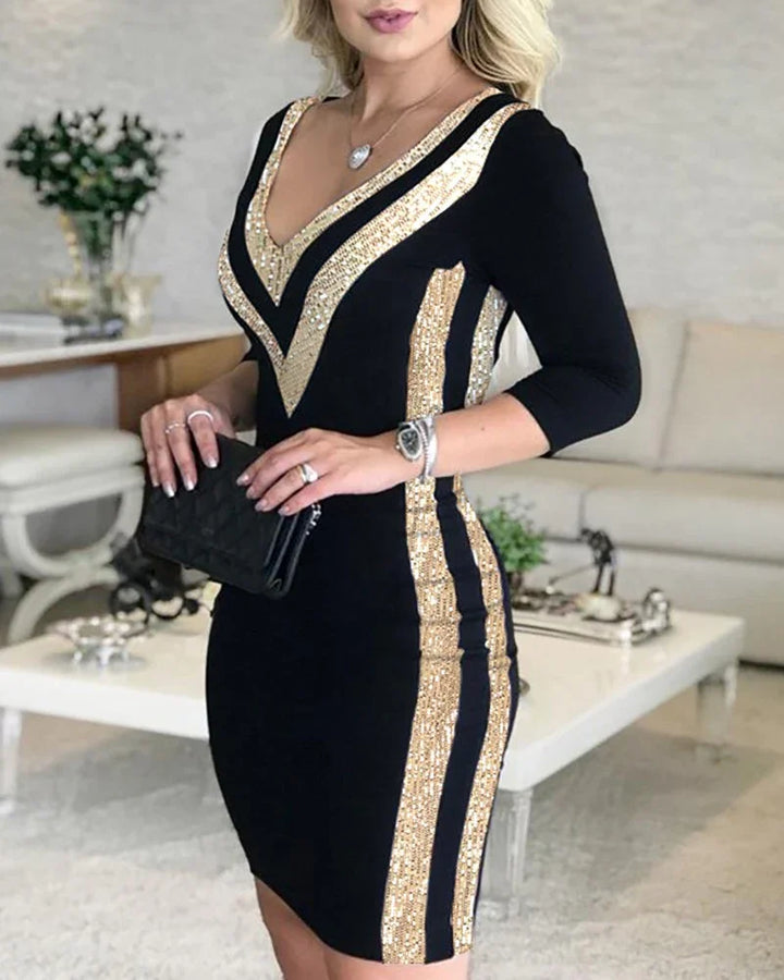 Woman wearing black bodycon dress with gold sequin V-neck and side stripes holding black clutch in living room