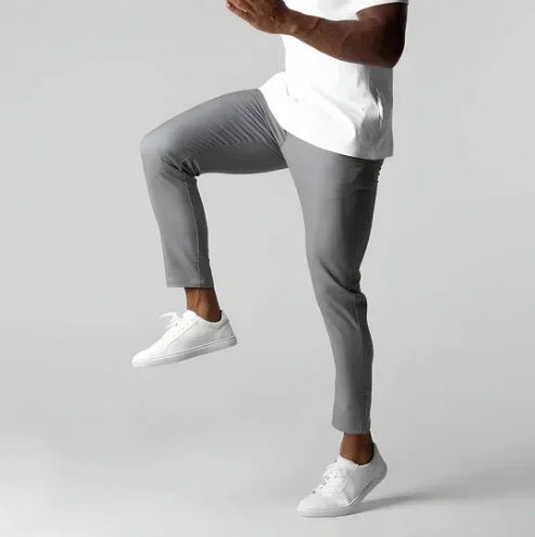 Man wearing gray slim-fit pants and white sneakers lifting one knee against gray background