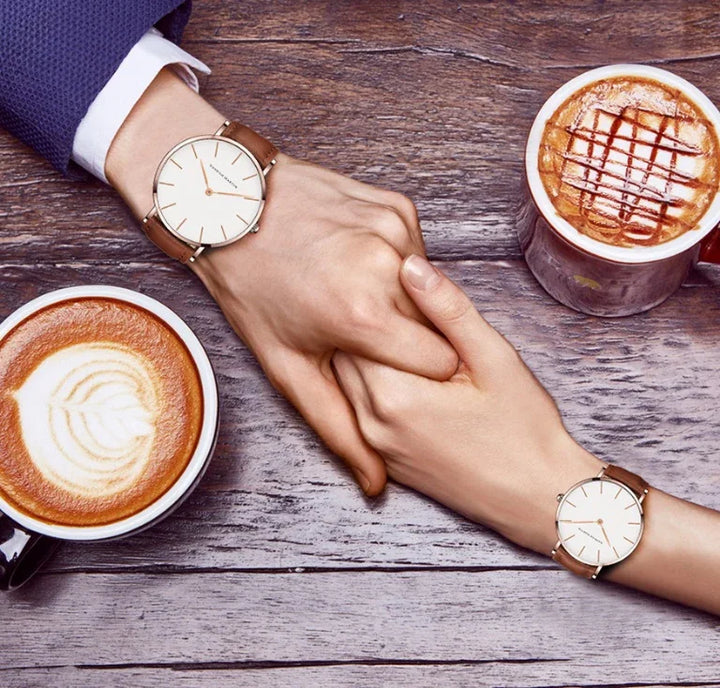Couple holding hands on wooden table wearing matching minimalist watches with brown leather straps and drinking coffee