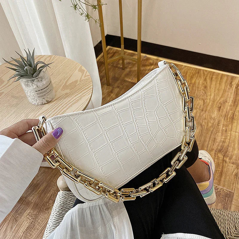 White crocodile texture handbag with gold chain strap held by hand over black pants and wooden floor