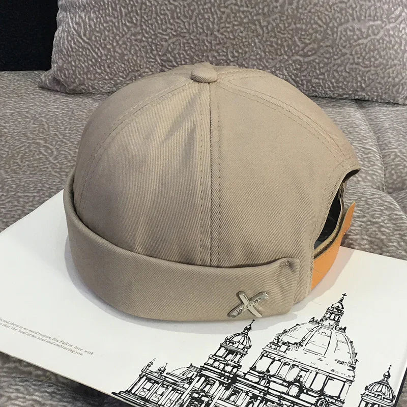 Beige cotton beanie hat with metal pin on side, placed on book with architectural sketches