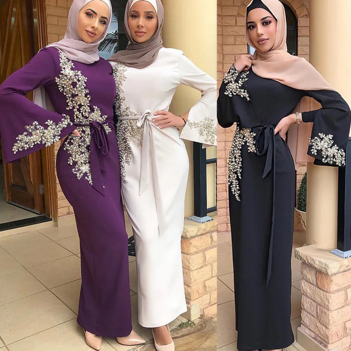Three women wearing elegant long-sleeve abaya dresses with floral bead embroidery and belted waist, in purple, white, and black colors, paired with hijabs