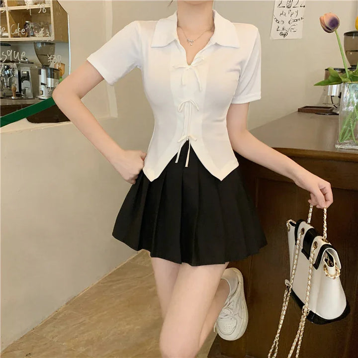 Woman wearing white short-sleeve top and black pleated skirt holding white handbag with gold chain in cafe