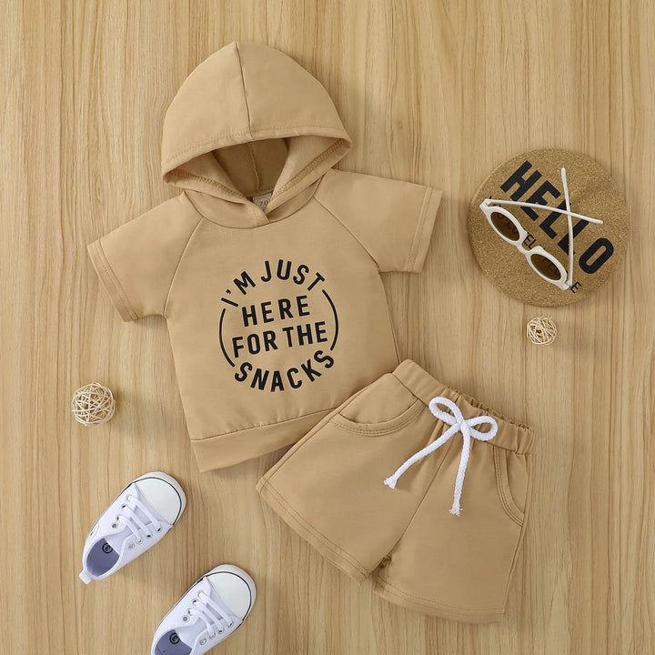 Beige toddler hoodie with "I'm just here for the snacks" text and matching shorts, white sneakers, sunglasses