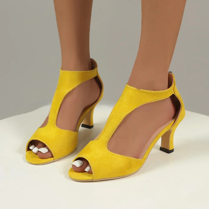 Yellow suede open-toe high heel sandals on feet with white pedicure against neutral background