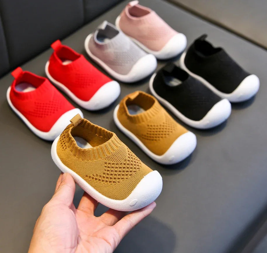 Hand holding mustard baby shoes with white soles, surrounded by similar shoes in red, pink, gray, and black
