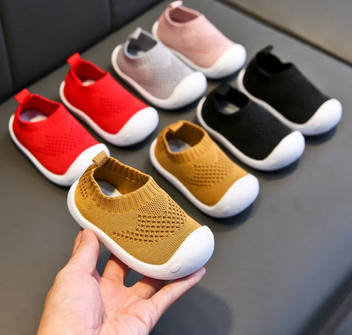Hand holding mustard baby shoes with white soles, surrounded by similar shoes in red, pink, gray, and black