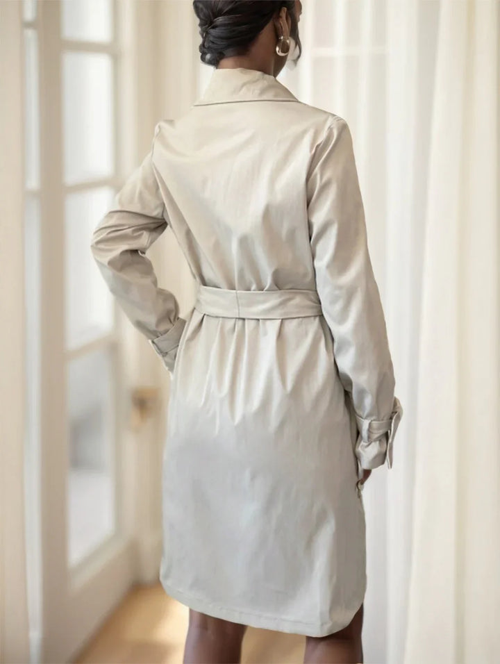 Back view of woman wearing beige belted trench coat with long sleeves in bright indoor setting