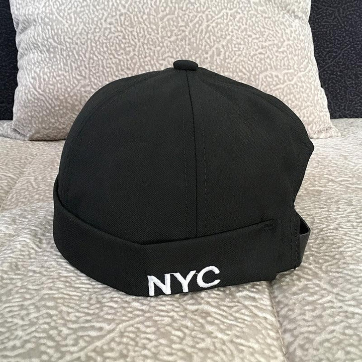 Black beanie cap with white NYC embroidery, resting on a beige patterned fabric surface