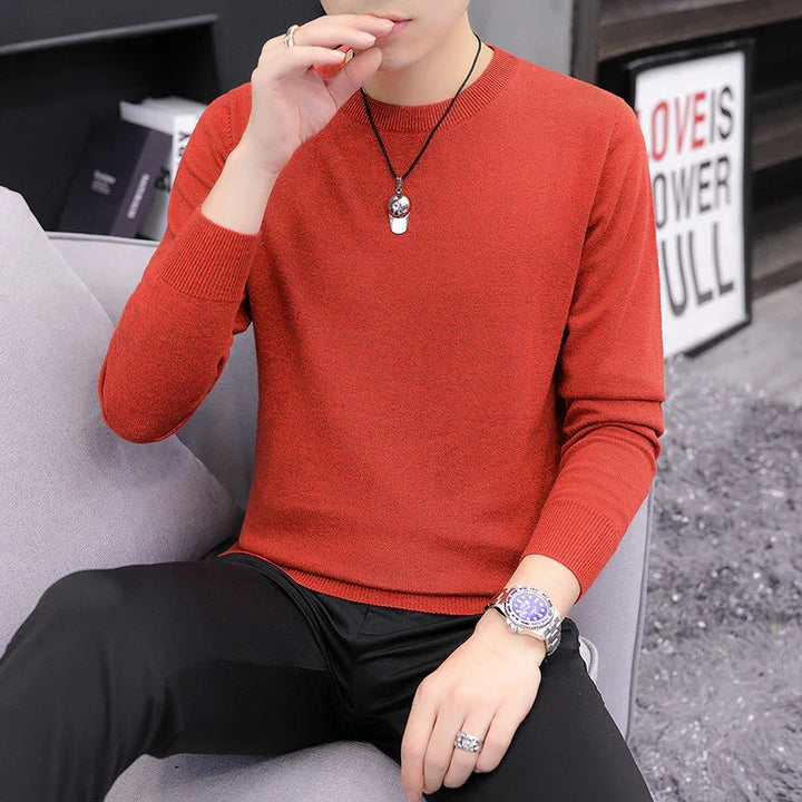 Man wearing red sweater and black pants sitting on gray couch with watch, rings, and pendant necklace