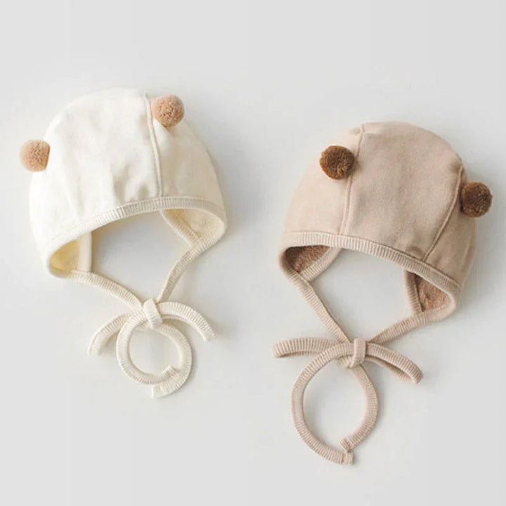 Two baby knit hats with pom-pom ears and ties in beige and cream colors on white background
