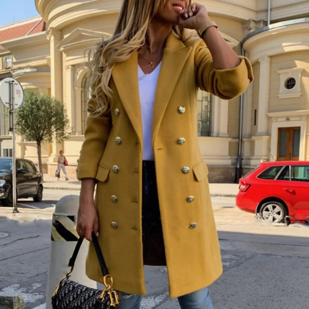 Woman wearing mustard double-breasted coat and holding black handbag in urban street setting