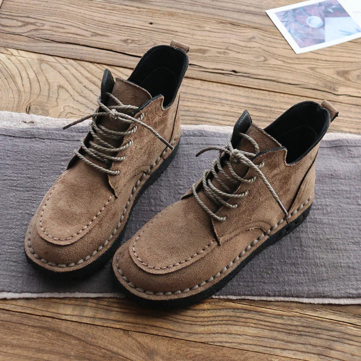 Brown suede casual lace-up ankle boots with stitched detailing on wooden floor and gray fabric