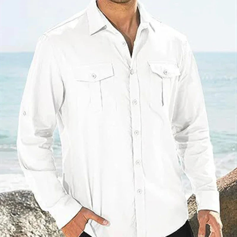Man wearing a white long-sleeve button-up shirt with front pockets standing by the ocean