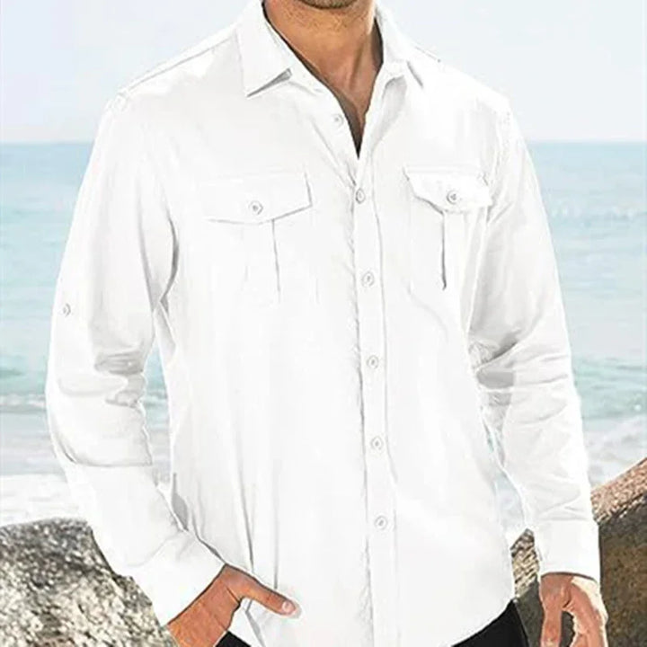 Man wearing a white long-sleeve button-up shirt with front pockets standing by the ocean