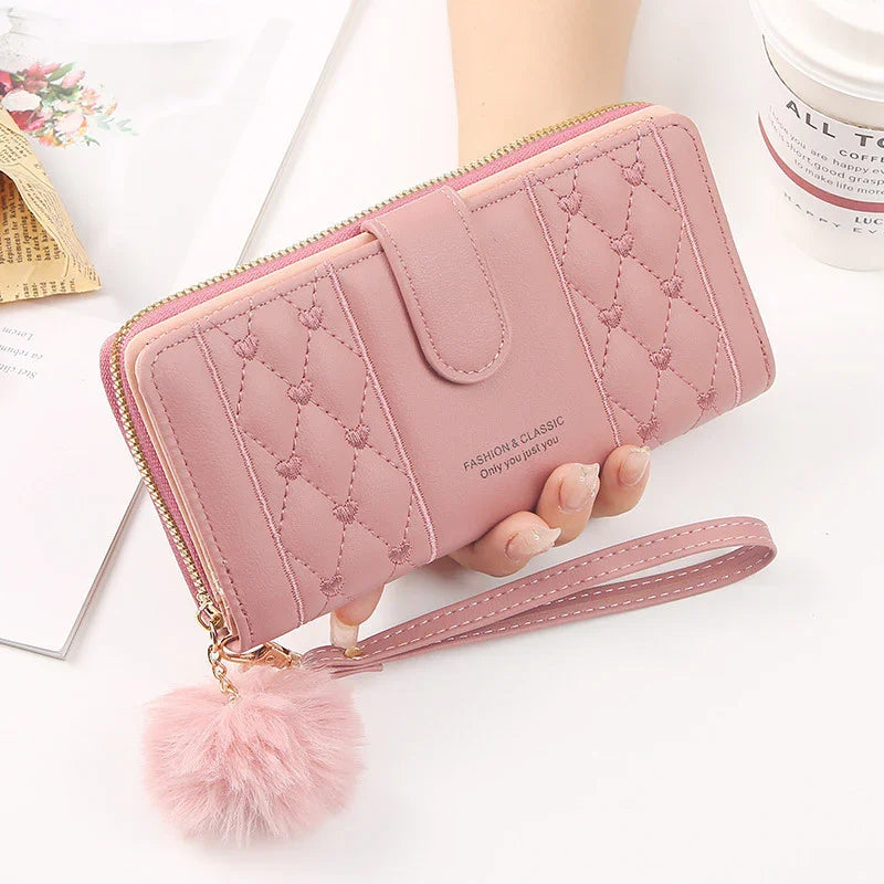 Pink quilted wallet with tassel pompom charm and wrist strap held by hand on white surface